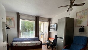 Quarto pequeno com cama, escrivaninha, cadeira, ventilador de teto e televisão.