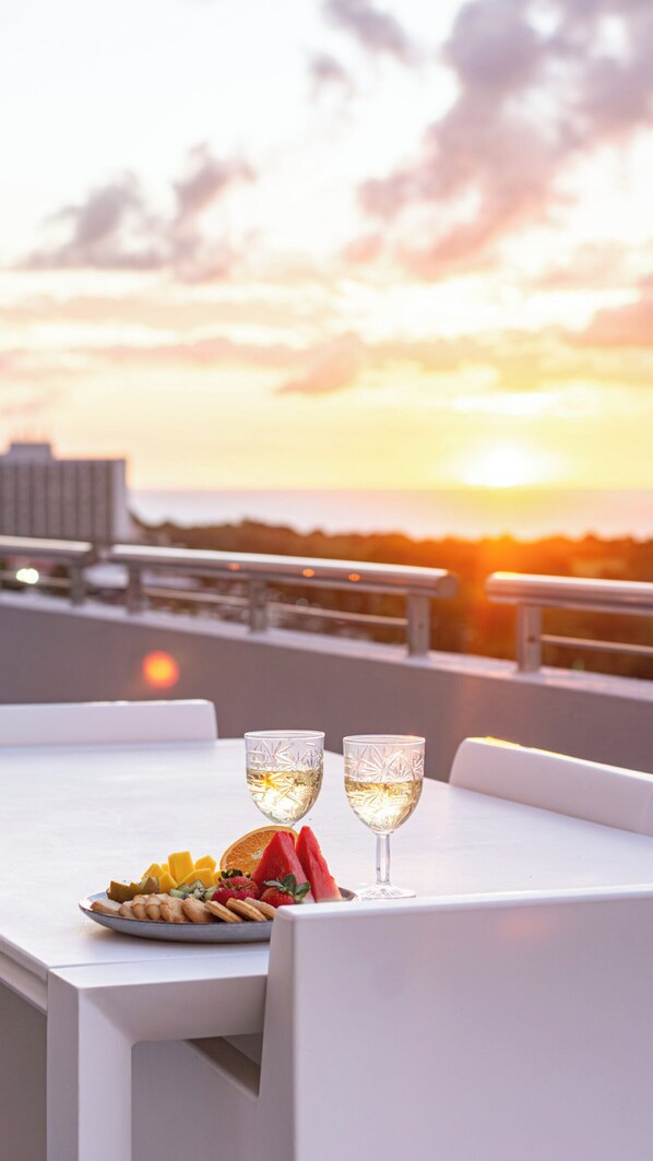 Een tafel met twee glazen witte wijn, een schaal fruit en uitzicht op de ondergaande zon boven de oceaan.