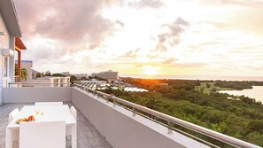 Een balkon met uitzicht op een zonsondergang boven het water, omringd door groen en gebouwen.