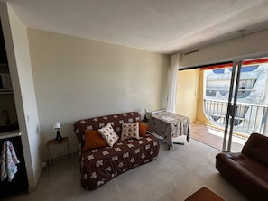 A living room with a sofa, a small table, and a chair. There is a balcony with a view of the outside. - Coeur de ville, studio avec parking N73 (La Grande-Motte)