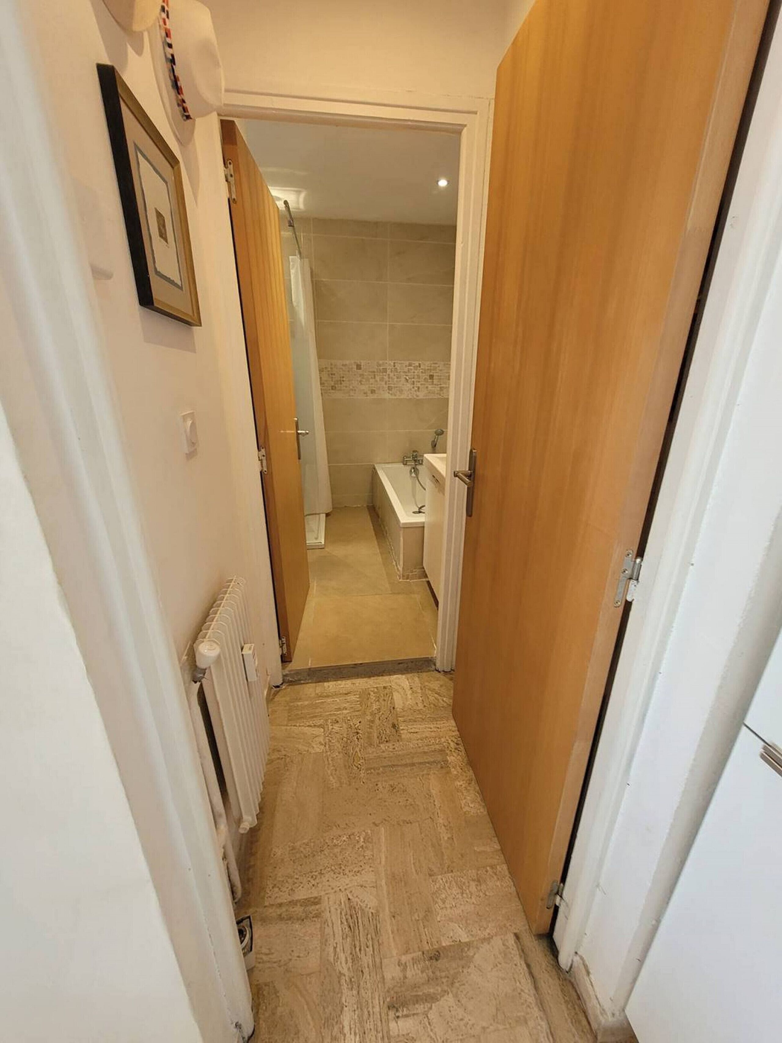 Un corridoio stretto con un termosifone a sinistra, un bagno con doccia e WC visibili attraverso una porta aperta, e una porta in legno a destra.