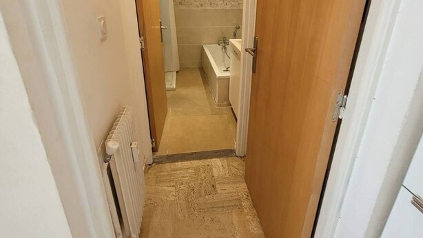 Un corridoio stretto con un termosifone a sinistra, un bagno con doccia e WC visibili attraverso una porta aperta, e una porta in legno a destra.