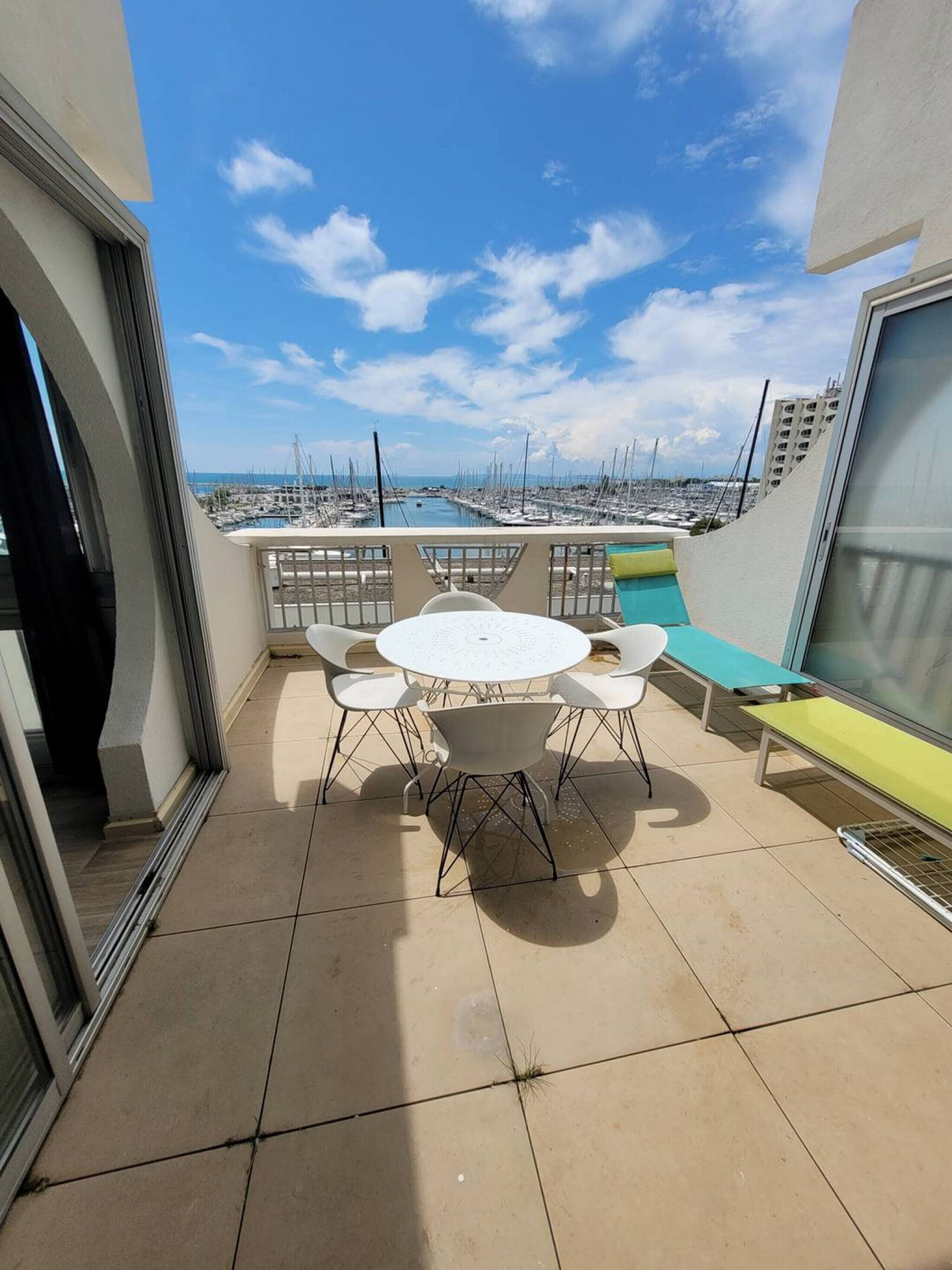 Un balcone con un tavolo rotondo e sedie, una panchina e vista su un porto turistico con barche.