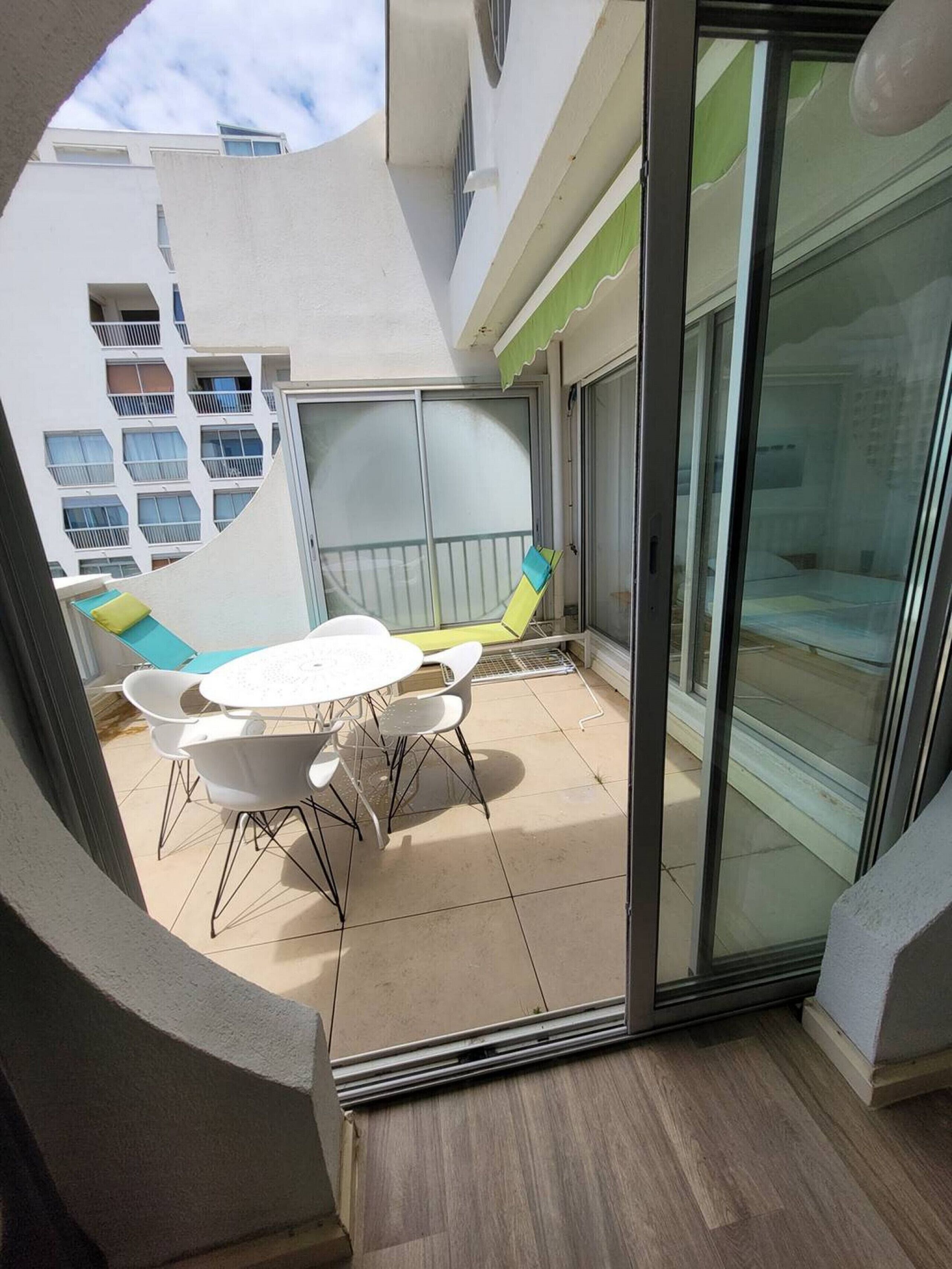 Un balcone con una porta vetrata, un tavolo bianco con sedie e vista su un edificio con numerose finestre.
