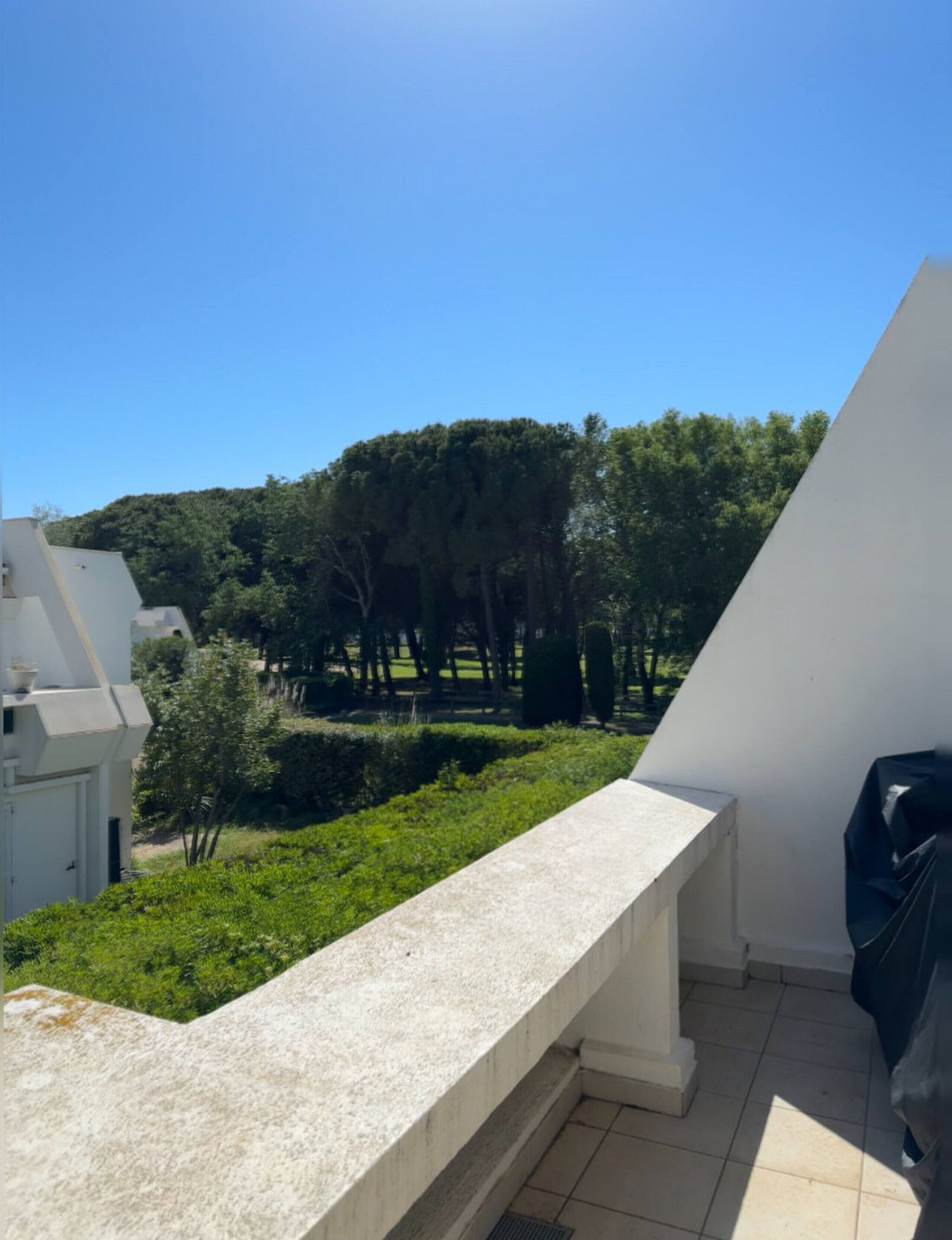 A balcony with a view of a green garden and trees, a white building, and a tiled floor.