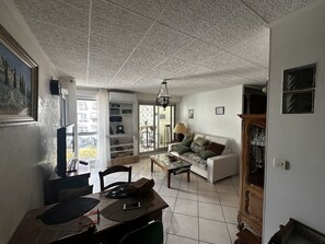 A living room with a sofa, a dining table with chairs, a coffee table, a bookshelf, and a large window. - Le temple du soleil, proche plage N76 (La Grande-Motte)