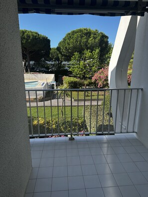 A balcony with a view of a swimming pool, greenery, and a white building.
