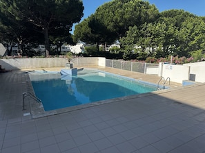 A rectangular swimming pool with a tiled deck, surrounded by a low white wall and trees. - Appartement pour 4, piscine et parking N75 (La Grande-Motte)