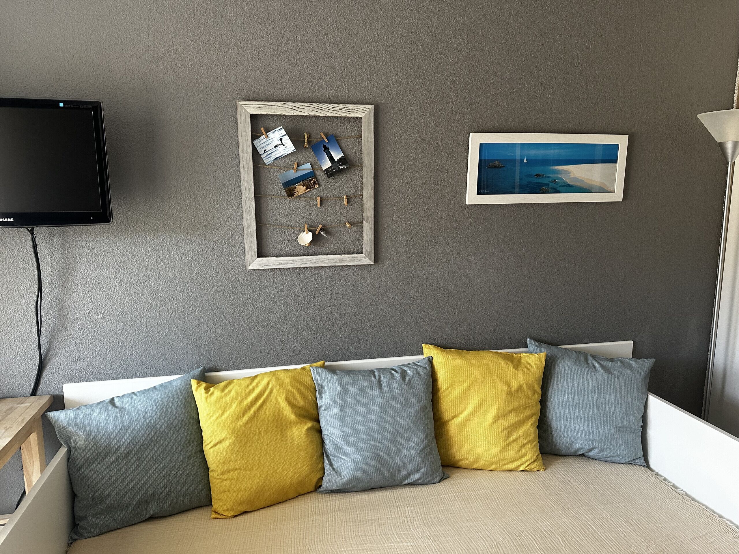 Una habitación con pared gris texturizada, un televisor de pantalla plana montado sobre un sofá blanco con cojines azules y amarillos, y dos cuadros enmarcados.