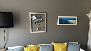 Ett rum med en grå, strukturerad vägg, en platt-TV som hänger ovanför en vit soffa med blå och gula kuddar, och två inramade tavlor.