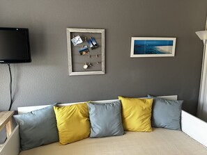 A room with a grey textured wall, a flat-screen TV mounted above a white sofa with blue and yellow cushions, and two framed pictures. - Appartement pour 4, piscine et parking N75 (La Grande-Motte)