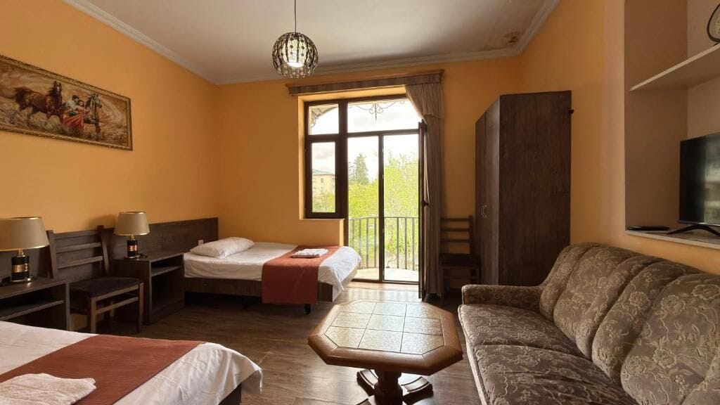 A hotel room with two beds, a sofa, a table, a TV, and a balcony with a view.