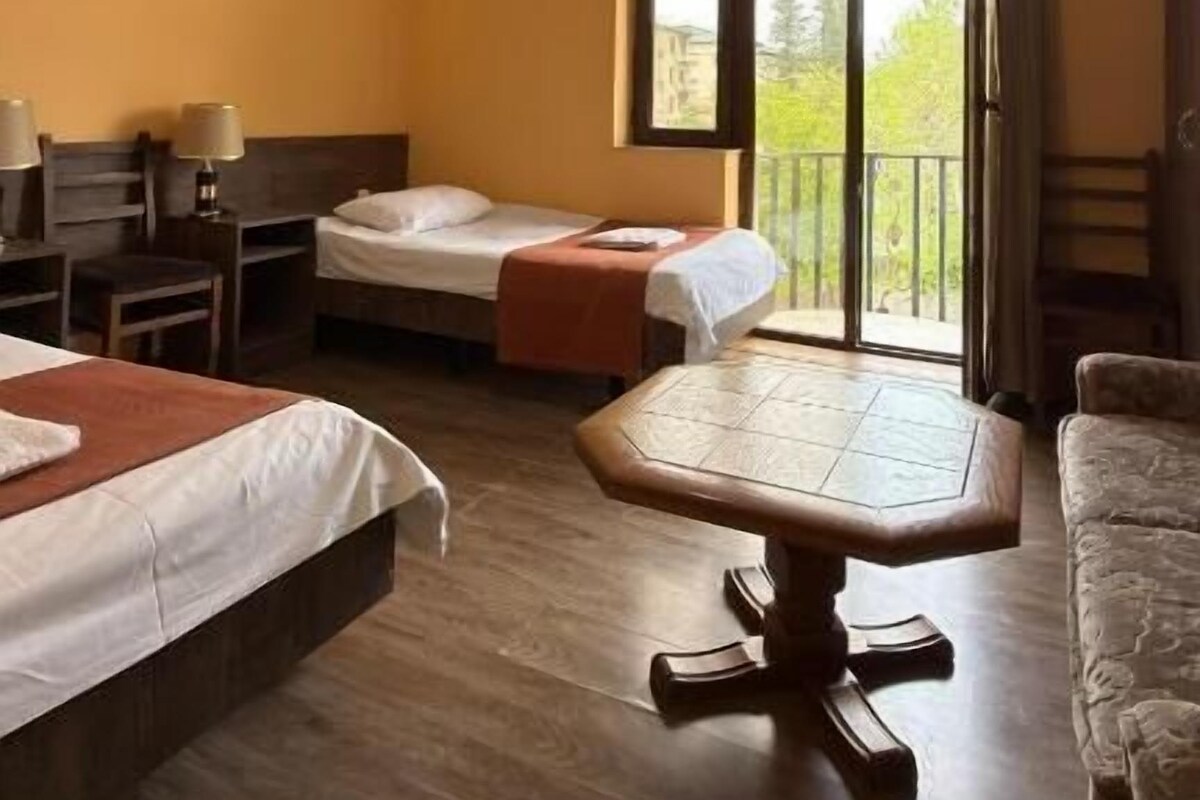 A hotel room with two beds, a dining table, a chair, a window with a view, and a chandelier.
