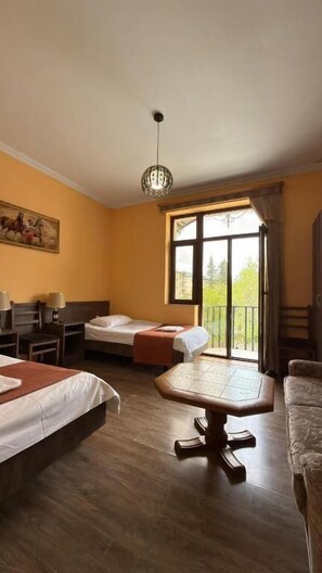 A hotel room with two beds, a dining table, a chair, a window with a view, and a chandelier. - Hotel Giorgi (Tskaltubo)