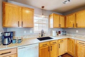 A kitchen with wooden cabinets, a dishwasher, a coffee maker, and a window. - The Brick Beauty | Dog-Friendly & Fenced Yard (Bentonville)
