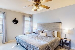 A bedroom with a large bed, a wooden headboard, two bedside tables with lamps, a ceiling fan, and a window with curtains. - The Brick Beauty | Dog-Friendly & Fenced Yard (Bentonville)