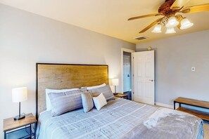 Ein Schlafzimmer mit einem Holz-Kopfteil, einem Bett mit vielen Kissen, einem Nachttisch mit Lampe, einem Deckenventilator und einer Tür im Hintergrund.