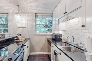 Una cocina con un lavamanos doble, estufa y microondas. Hay armarios y una ventana con vista a árboles.