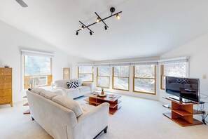 A spacious living room with a sofa, coffee table, TV, and large windows. - Lakefront 6BR with infinity pool, sauna & kayaks (Rogers)