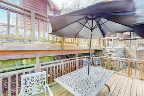 A wooden deck with a table and chairs, an umbrella, and a stone wall. - Lakefront 6BR with infinity pool, sauna & kayaks (Rogers)