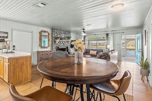 A spacious living area with a round wooden table, comfortable seating, a stone fireplace, and a view through large windows. - 2BR Sunroom Charm & Lakefront Dock Living (Hot Springs)