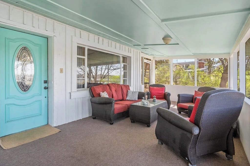 Une véranda couverte avec une porte de couleur turquoise, un canapé noir avec des coussins rouges et une table basse en verre.