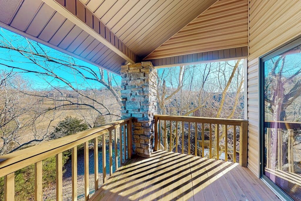 A wooden deck with a stone fireplace, large windows, and a view of the surrounding landscape.