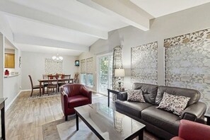 A modern living room with a leather sofa, a glass coffee table, and a dining area with a wooden table and chairs. - 3BR Inviting woodland retreat - direct bike trail (Bella Vista)