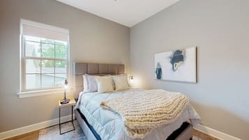 A bedroom with a bed, a bedside table, a window, and a wall-mounted artwork.