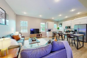 Ein modernes Wohnzimmer mit Sofa, Couchtisch und Fernseher. Angrenzend befindet sich eine Küche mit weißen Schränken, ein Essbereich mit Stühlen und ein Kühlschrank.
