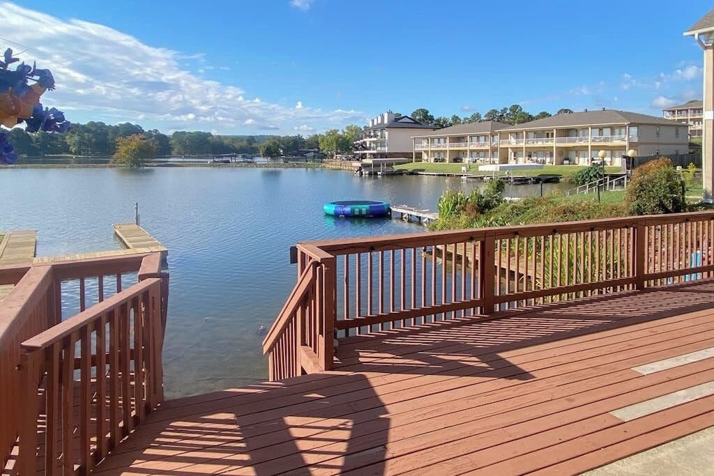 A wooden deck with a view of a lake, a floating dock, and a building with multiple balconies.