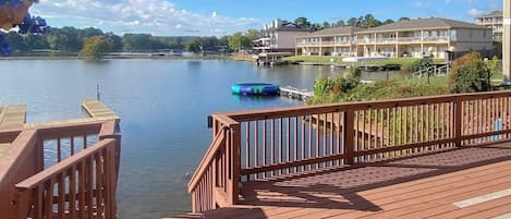 A wooden deck with a view of a lake, a floating dock, and a building with multiple balconies.