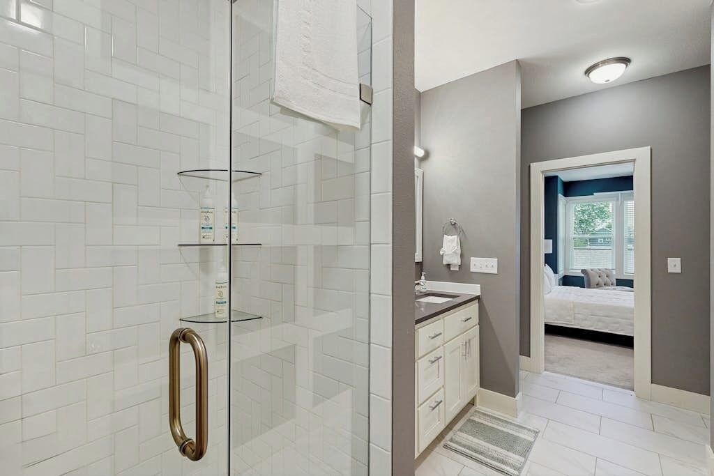A modern bathroom with a glass shower enclosure, white tiled walls, a vanity with a sink, and a visible bedroom with a bed and window.
