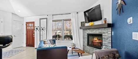 A living room with a fireplace, a television, and a view of the outside.