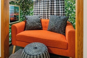 A room with a sofa, two pillows, a round ottoman, and a framed picture on the wall. - Spacious 4BR - Firepit - Garage Parking - W&D (Bentonville)
