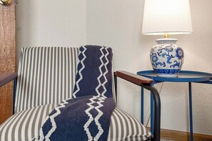 A chair with a striped cushion and a patterned throw blanket, a blue and white vase lamp on a small table beside it, and a wooden door in the background. - Spacious 4BR - Firepit - Garage Parking - W&D (Bentonville)