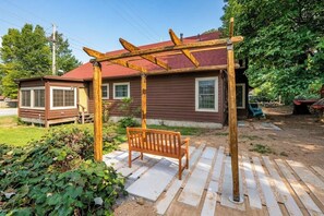 Une pergola en bois avec un banc, une maison avec un toit rouge et un jardin agrémenté de pierres plates servant de pas.