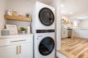 A laundry area with two stacked front-loading washing machines, a wooden shelf with jars, a framed certificate, and a potted plant. - B Street Bungalow | Bike-In/Bike-Out & Game Room (Bentonville)