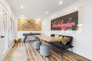A modern living room with a wooden floor, a black sofa with cushions, a wooden coffee table, a bicycle mounted on the wall, and a neon sign. - B Street Bungalow | Bike-In/Bike-Out & Game Room (Bentonville)