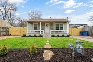 Une maison de plain-pied avec un porche à l’avant, un petit jardin devant la maison avec un chemin bordé de pierres, et un compteur d’eau ou d’électricité visible au premier plan.