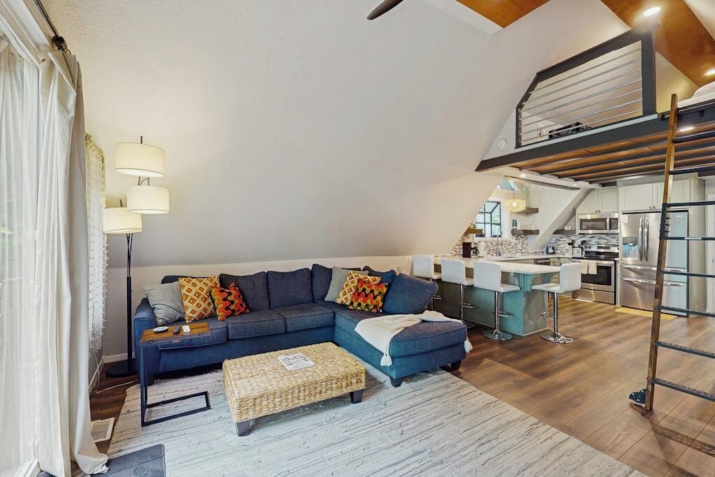 Una sala de estar moderna con un sofá seccional, un área de cocina y un espacio tipo loft con una escalera.