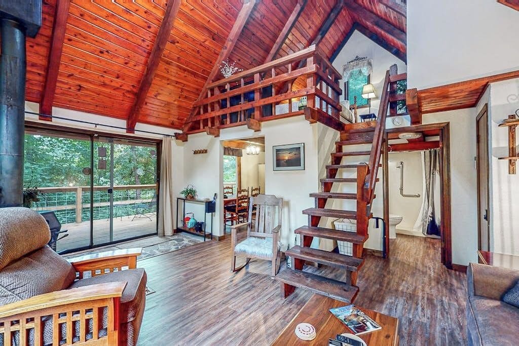 A spacious living area with a wooden staircase leading to an upper level, a large window offering a view of greenery, and a cozy seating area with a fireplace.