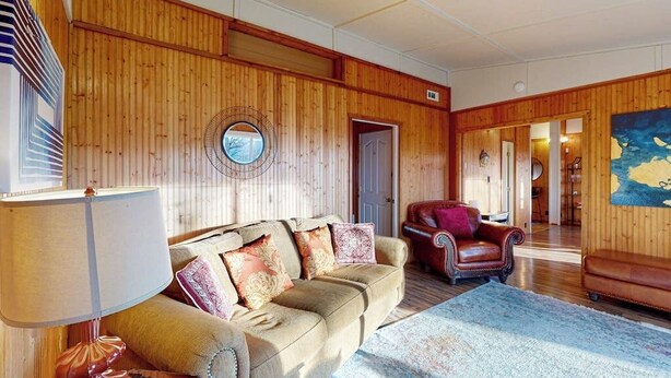 A room with a wooden paneled wall, a round mirror, a sofa with patterned cushions, a red leather armchair, and a large rug. - Quiet 2BR Lakeview Dog Friendly - Deck - Firepit (Eureka Springs)
