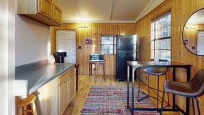 A compact kitchen with wooden walls, a refrigerator, a microwave, a coffee maker, and a dining area with a table and chairs. - Quiet 2BR Lakeview Dog Friendly - Deck - Firepit (Eureka Springs)