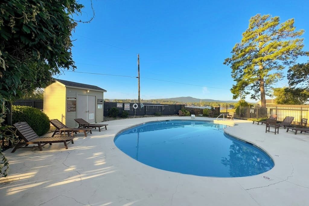 A swimming pool with lounge chairs and a small building, surrounded by trees and a clear sky.