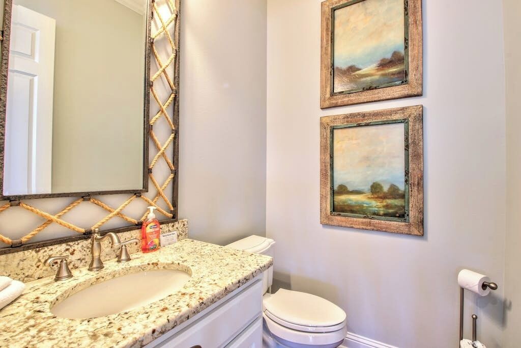Een badkamer met een wastafel met een granieten blad, een spiegel, een toilet en twee lijstwerken van landschappen aan de muur.