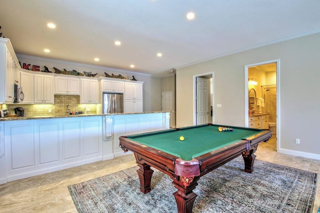 A pool table in a room with a kitchen area and a doorway leading to a bathroom.