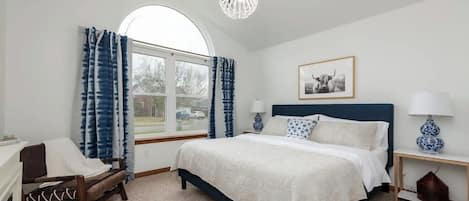 A bedroom with a bed, a chair, a nightstand, a lamp, a picture on the wall, and a window with curtains.