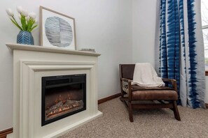 A fireplace with a fire, a framed artwork above it, a vase with flowers, and a chair with a blanket. - 4BR with private pool, patio & firepit (Bentonville)