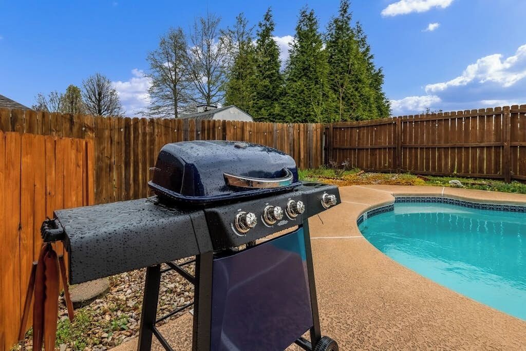A barbecue grill with a lid, a pool, and a wooden fence in the background.